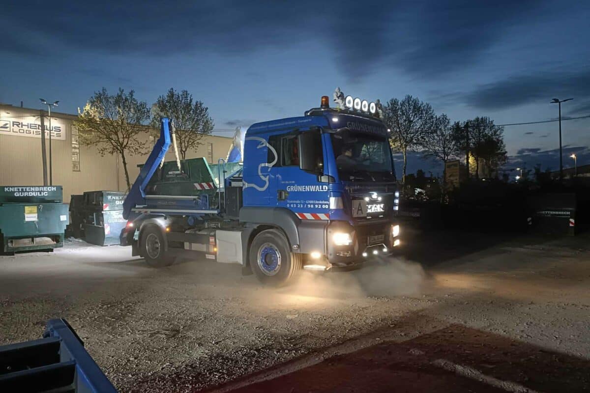 Ein blauer Schwerlast-Lkw mit eingeschalteten Scheinwerfern steht in der Abenddämmerung auf einem Gewerbegrundstück und ist bereit für seine Verkehrssicherungsaufgaben.